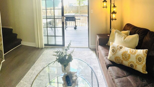 A living room with a glass coffee table, a brown leather sofa with patterned cushions, a sliding glass door leading to an outdoor patio, and two wall-mounted floor lamps.