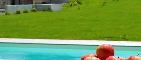 Zona junto a la piscina con un bol de manzanas, una terraza de madera y un edificio de piedra al fondo.