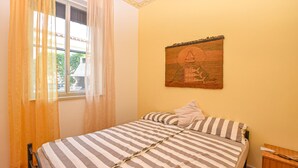 Un dormitorio con cama, ventana con cortinas, una obra de arte colgada en la pared, mesita de noche y una silla.