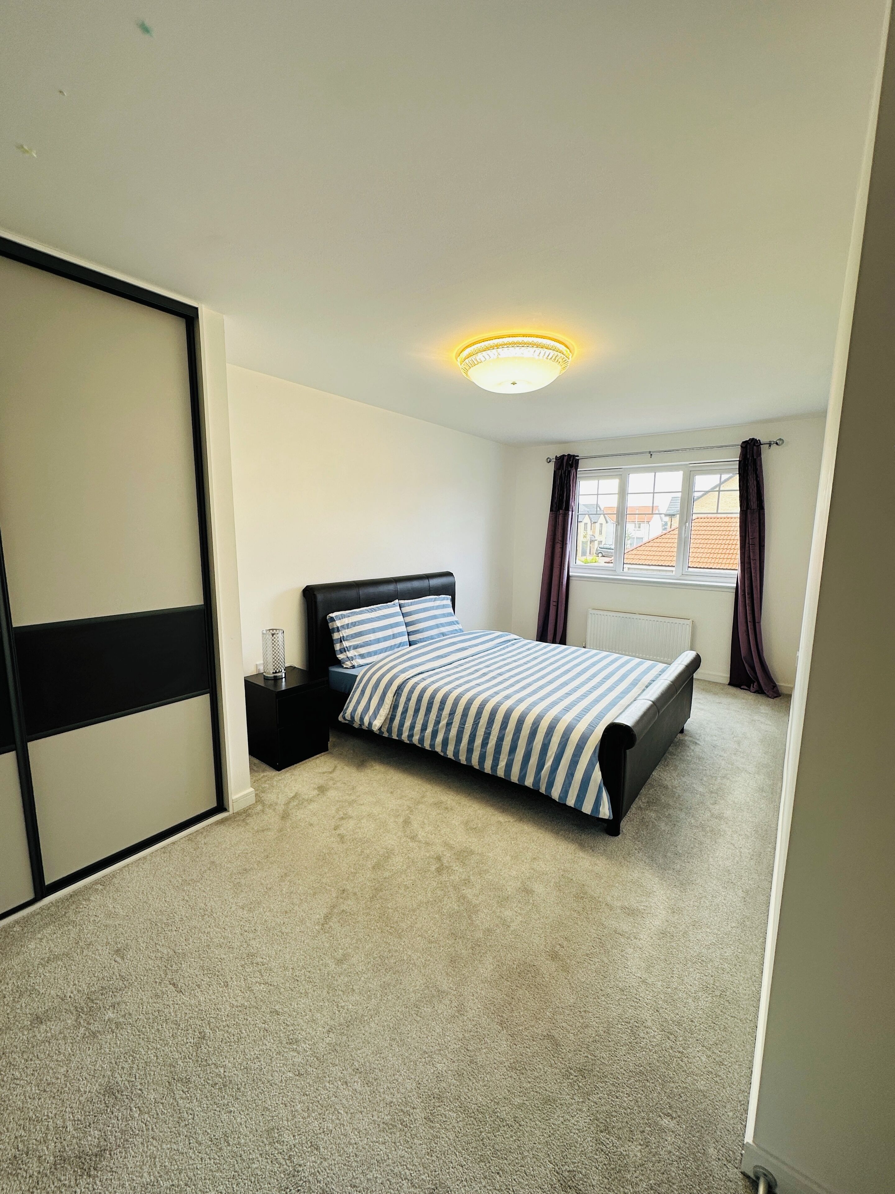 Un dormitorio con cama, mesita de noche, radiador y ventana con cortinas.