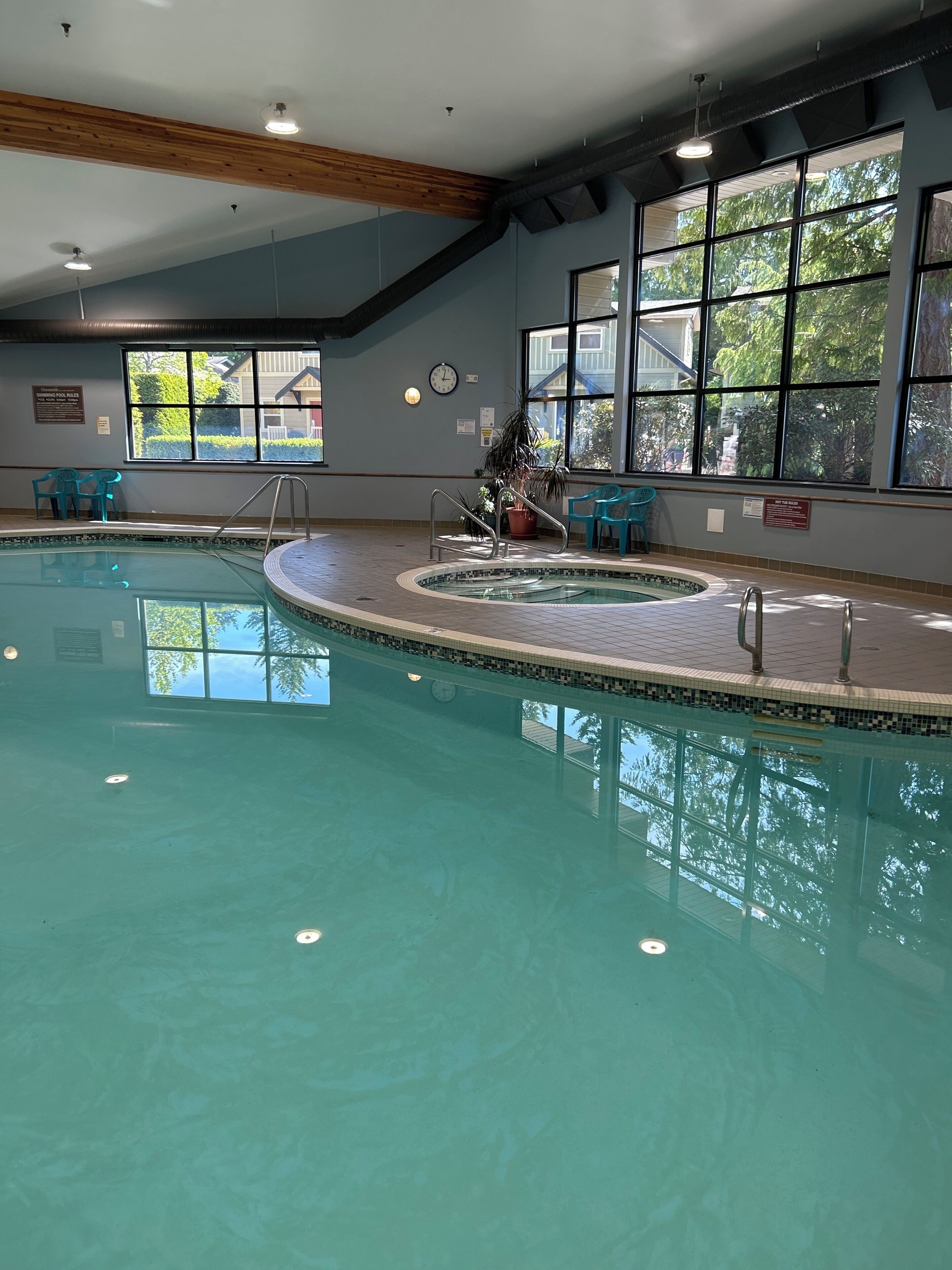 An indoor swimming pool with clear water, wooden beams on the ceiling, and large windows with a view of trees.