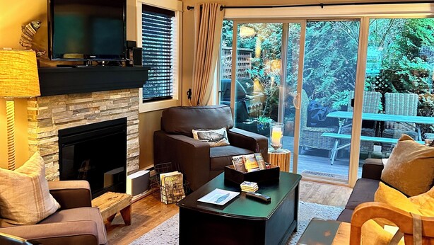 A cozy living room with a fireplace, comfortable seating, and a large window offering a view of the outdoors. - Moon shell Cottage-a relaxing cozy retreat for all seasons. (Parksville)