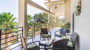 Un balcón con mobiliario de mimbre y plantas en macetas, con vistas a un jardín con palmeras.