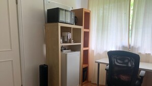 A compact room with a white desk, a black office chair, a microwave, a coffee maker, and a small refrigerator.