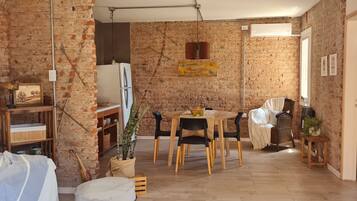 Una habitación acogedora con pared de ladrillo, una mesa de comedor de madera con sillas, un refrigerador y un pequeño rincón de descanso con una silla y una planta.