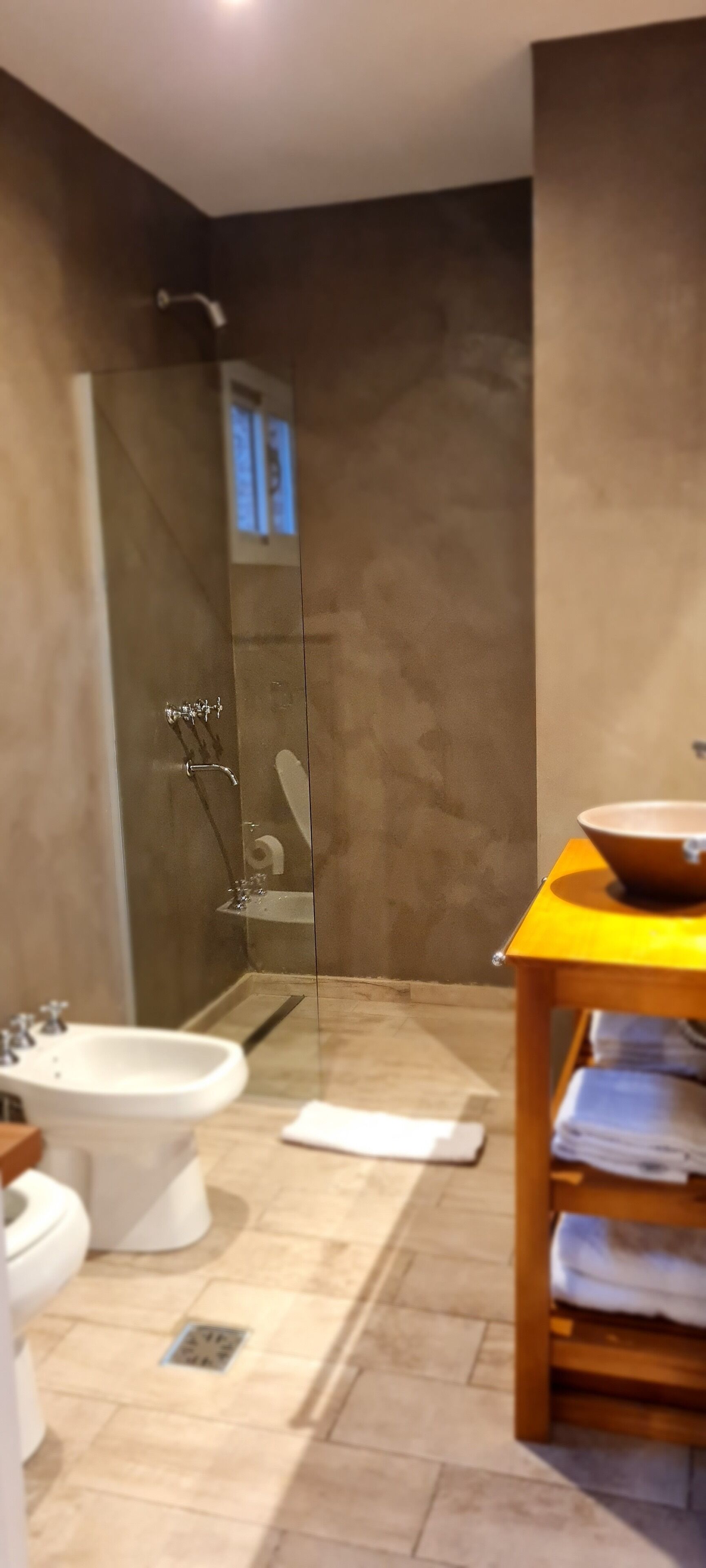 A modern bathroom with a glass-enclosed shower, a bidet, a sink, and a wooden vanity with towels.