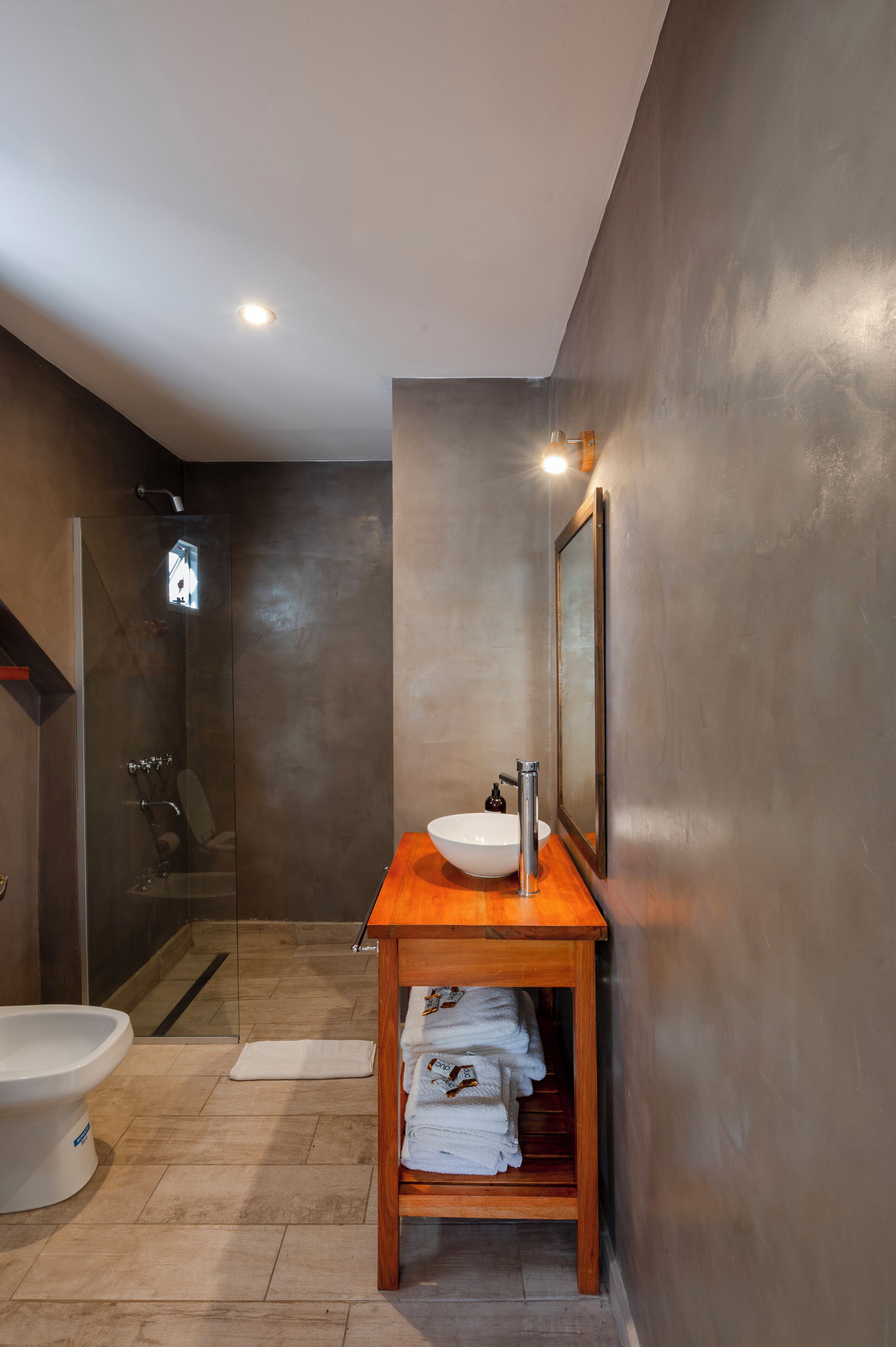 Un baño moderno con ducha envuelta en vidrio, tocador de madera con lavamanos blanco y espejo.