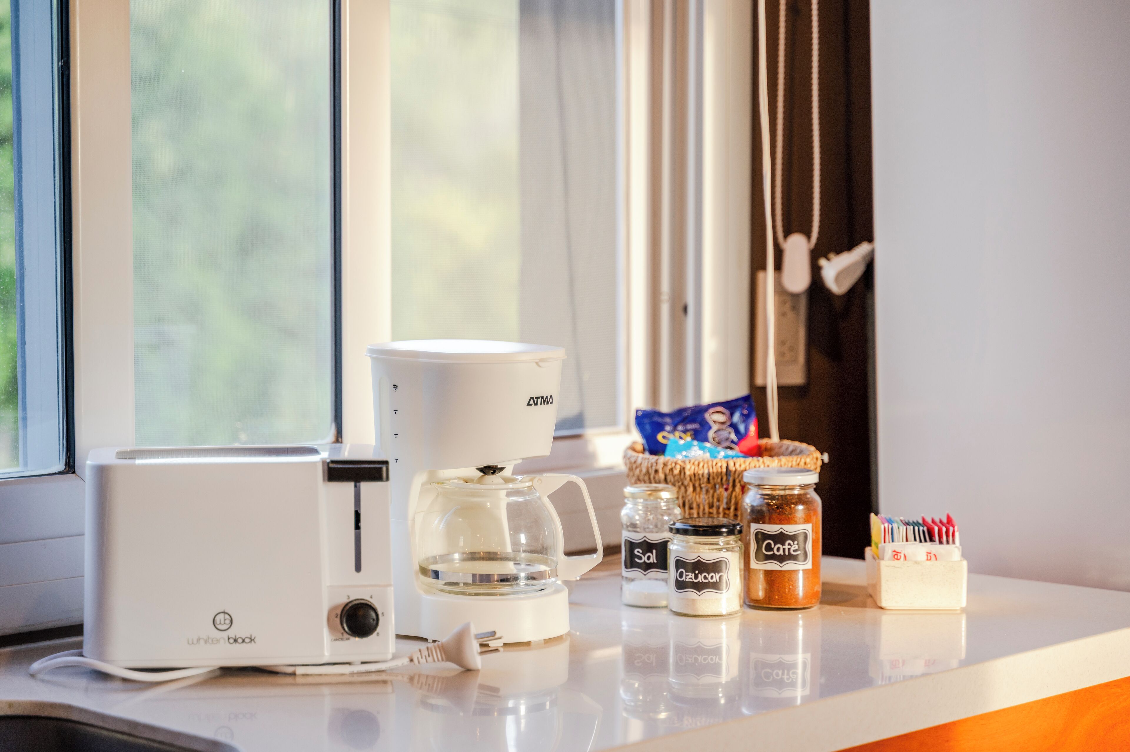 Una encimera de cocina con una tostadora blanca, una cafetera, botes de azúcar y sal, y una cesta de aperitivos.