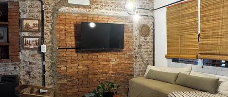 A living room with a brick wall, a flat-screen TV, a wooden coffee table, a sofa, and a window with blinds.