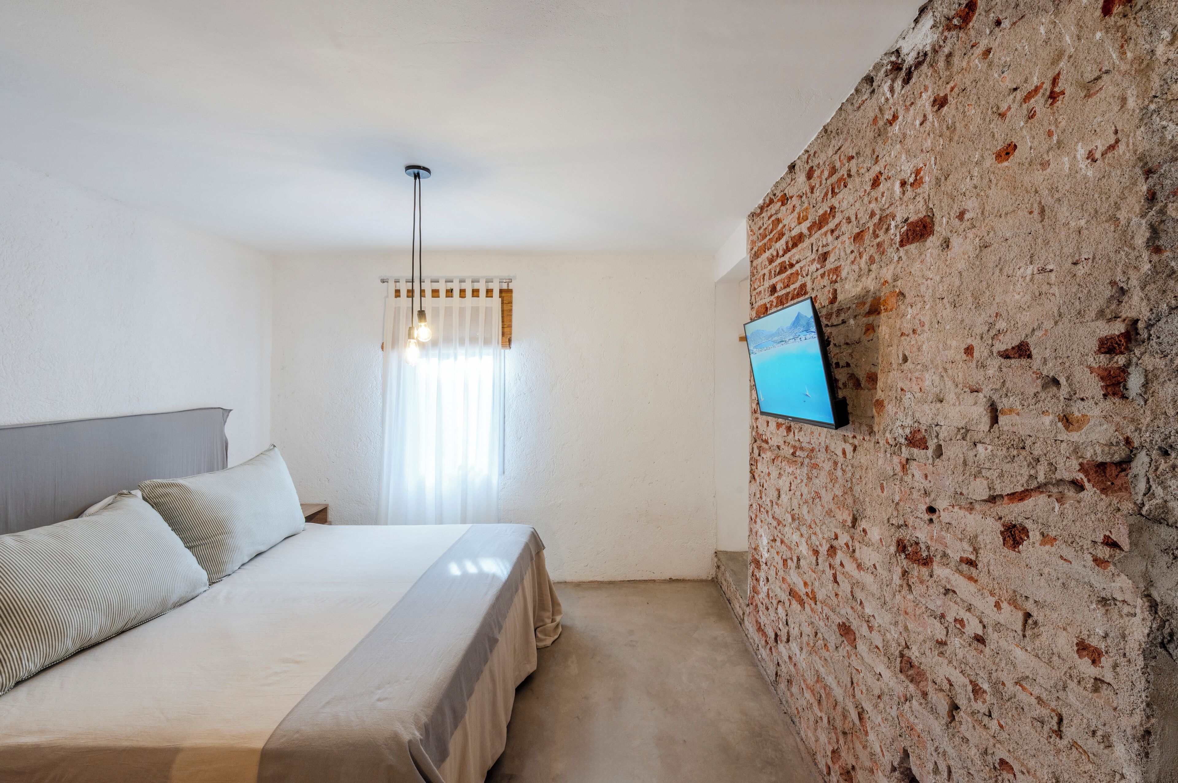 A bedroom with a bed, a brick wall, a TV mounted on the wall, and a window with curtains.