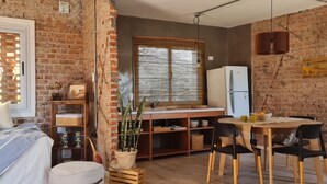 Habitación compacta con cama, kitchenette pequeño, zona de comedor y pared de ladrillo.