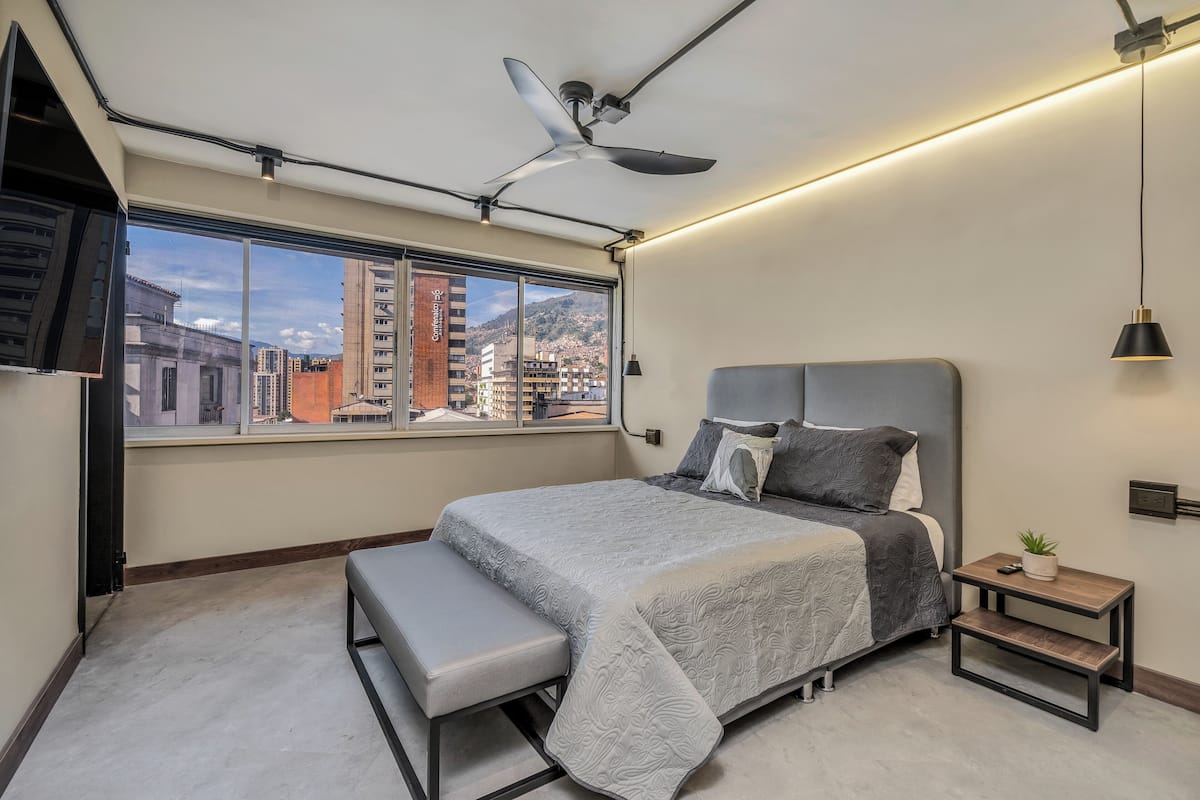 Een moderne slaapkamer met een groot bed, een bankje, een plafondventilator, een nachtkastje en een uitzicht op de stad door een groot raam.