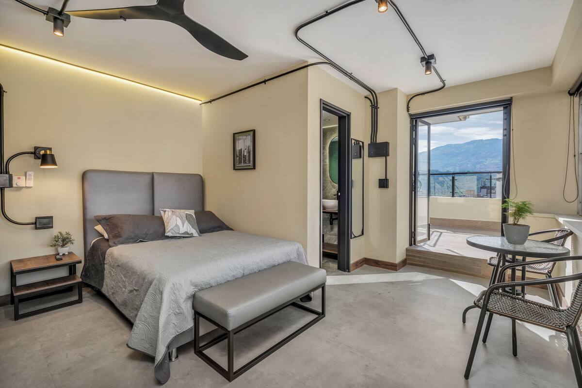 Een moderne slaapkamer met een bed, een bankje, nachtkastje en een balkon met een tafel en stoelen.