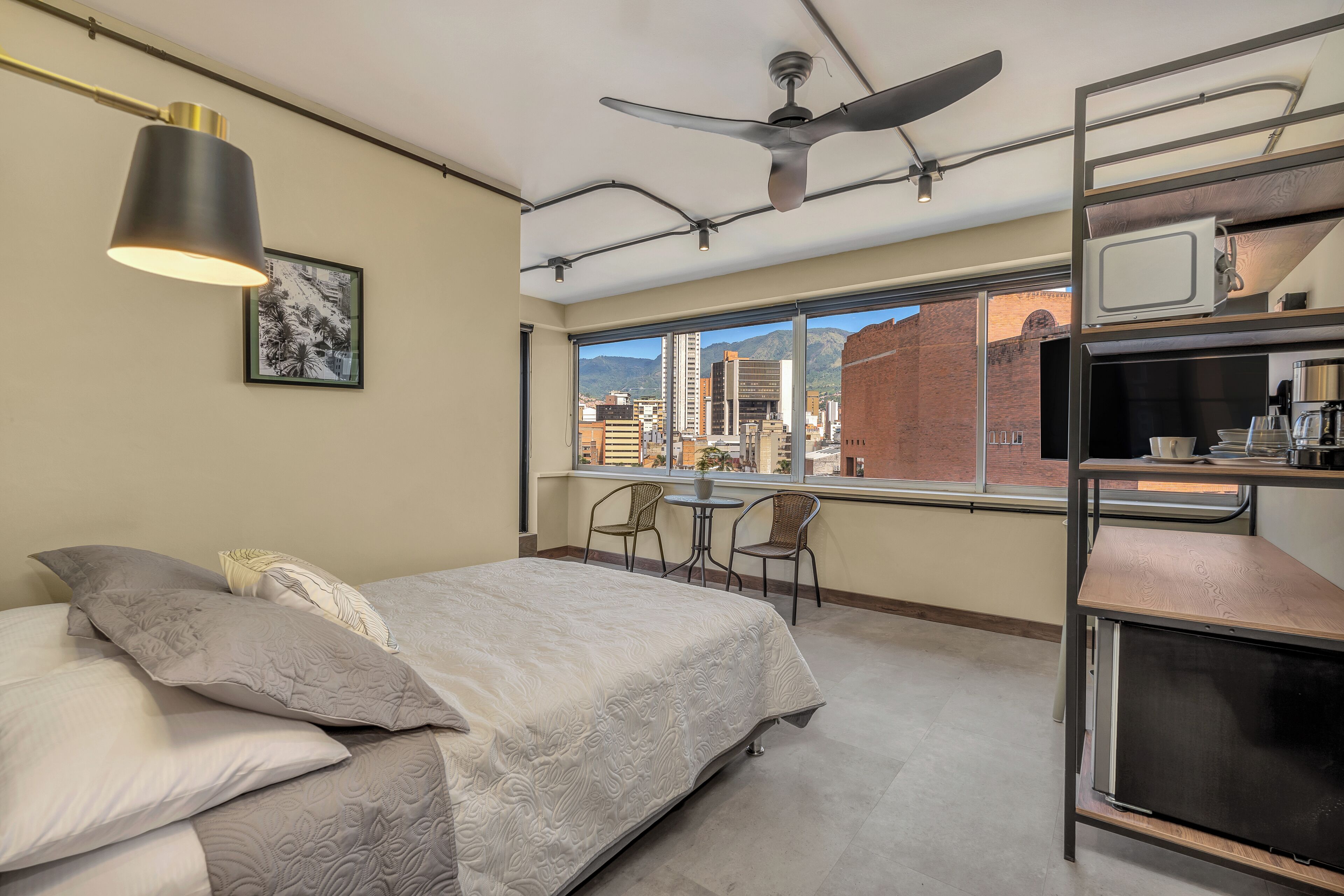 A bedroom with a bed, pillows, a ceiling fan, a window with a city view, and a desk with a chair.