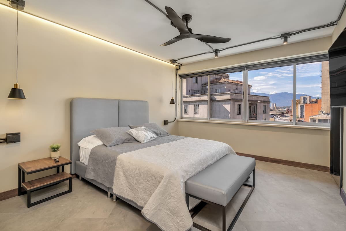 Een moderne slaapkamer met een bed, bankje, nachtkastje en een groot raam met uitzicht over de stad.