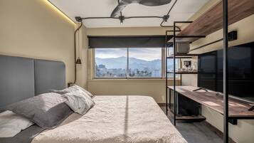 A modern bedroom with a large bed, a ceiling fan, a city view, and a kitchenette area.