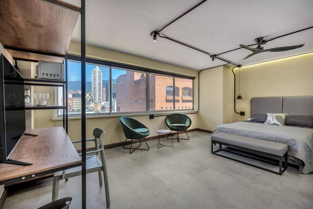 Een moderne hotelkamer met een groot raam, een bed, een bureau, twee stoelen en een plafondventilator.