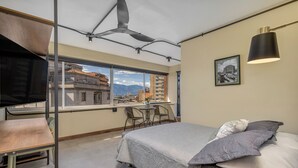 Ein Schlafzimmer mit einem Bett, einem Schreibtisch und einem Sessel, nahe eines Fensters mit Blick auf die Stadt.