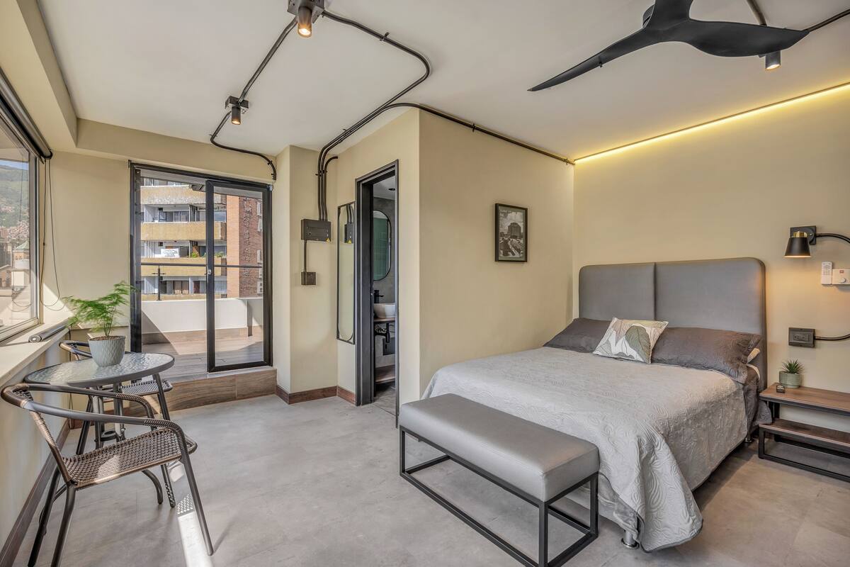Een moderne slaapkamer met een groot bed, een bankje, een kleine tafel met stoelen en een balkon met uitzicht op gebouwen.