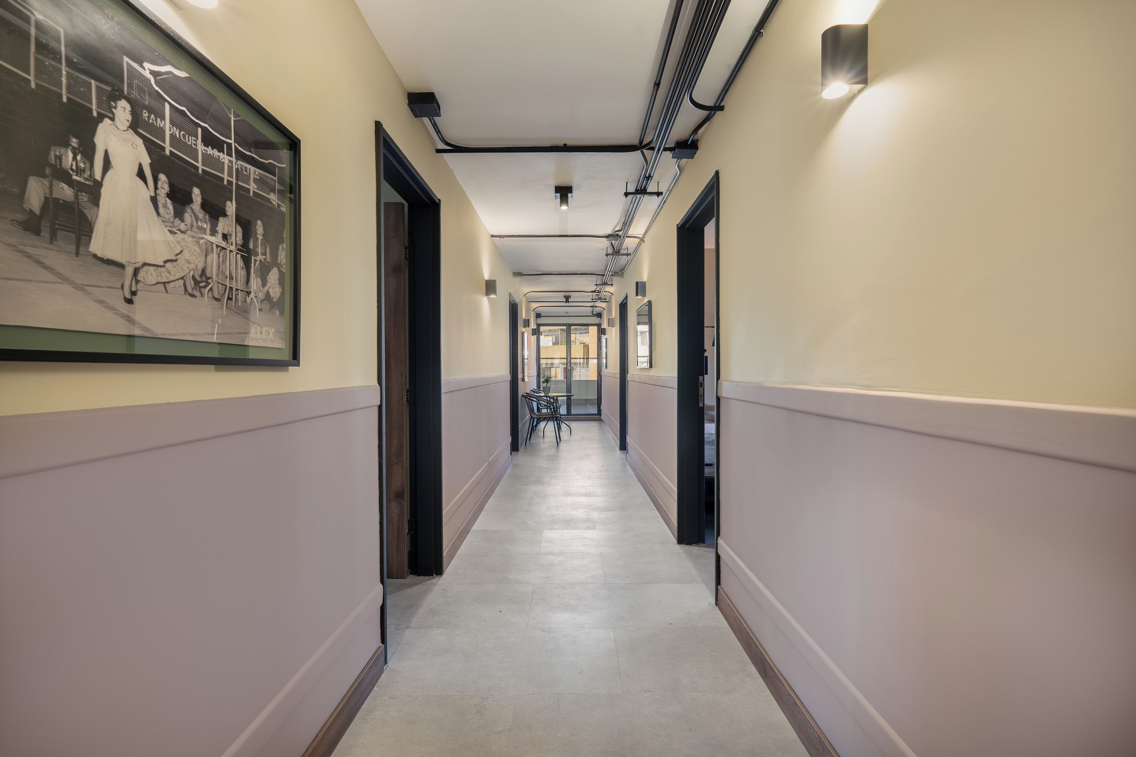 A hallway with a framed historical photograph on the left, a long corridor with doors on both sides, and a ceiling with exposed pipes and lighting fixtures.