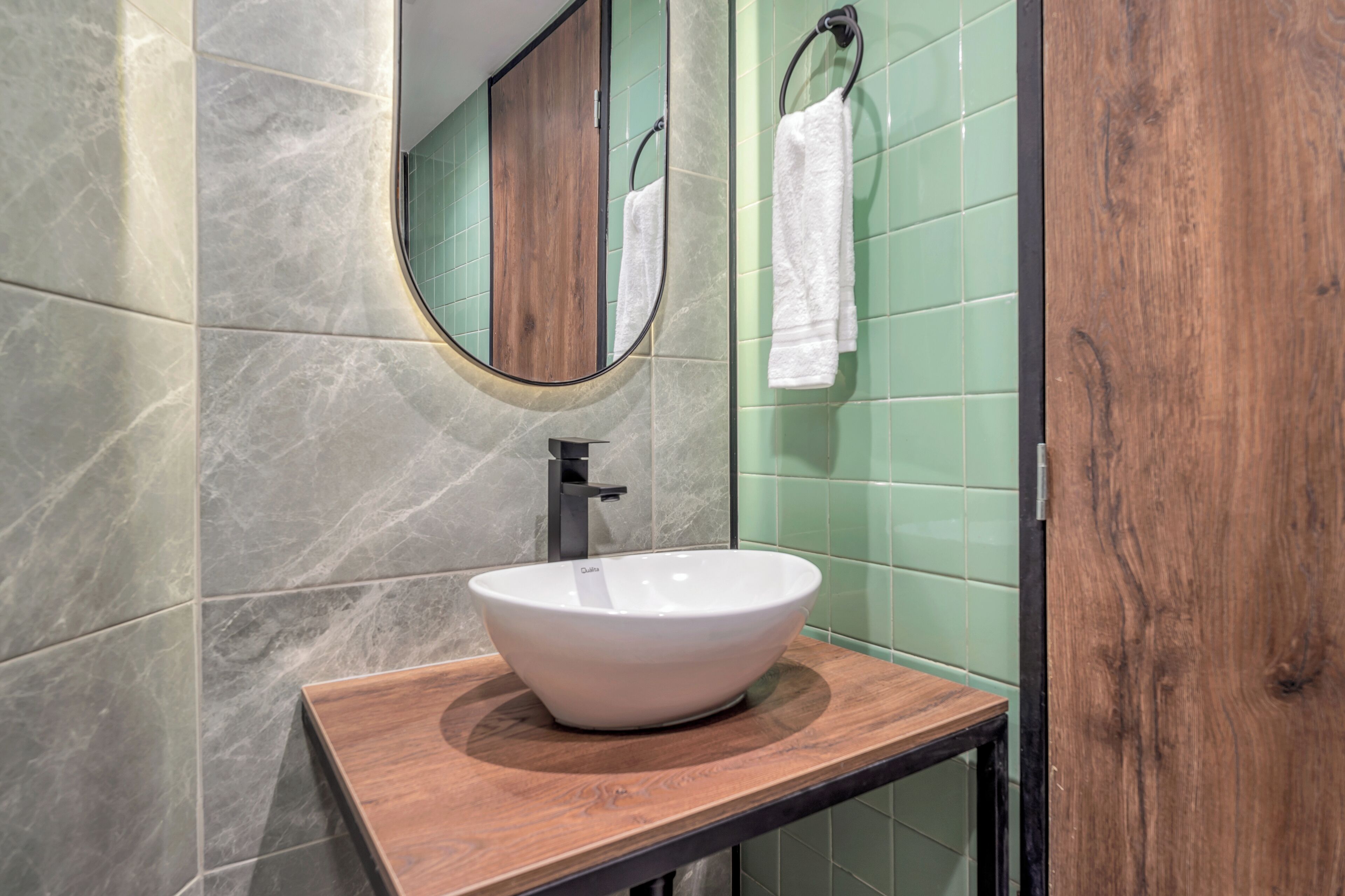 A bathroom with a wooden vanity, a white vessel sink, a mirror, and green tiled walls.