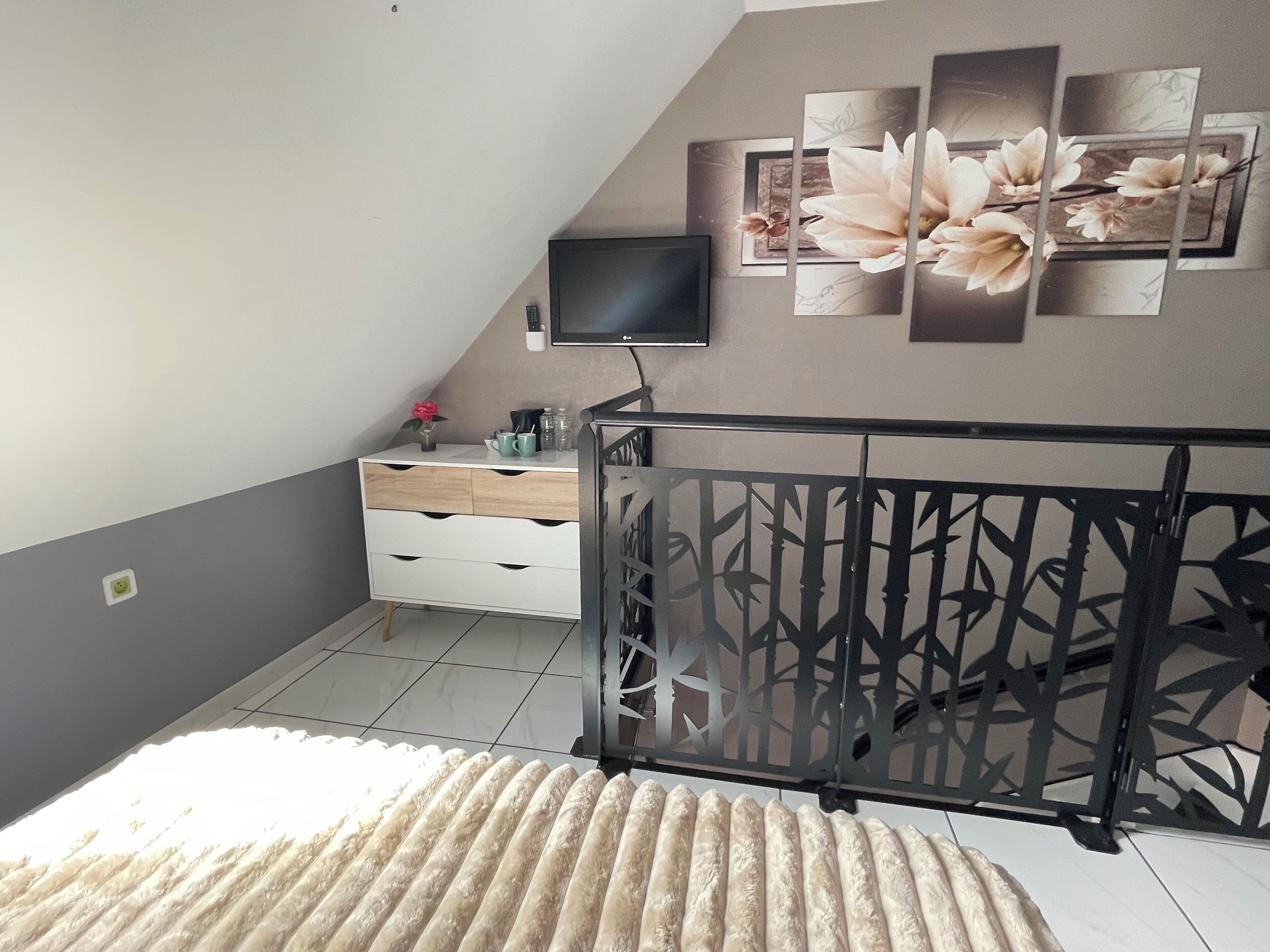 A bedroom with a sloped ceiling, a TV mounted on the wall, a white dresser, and a decorative wall panel.