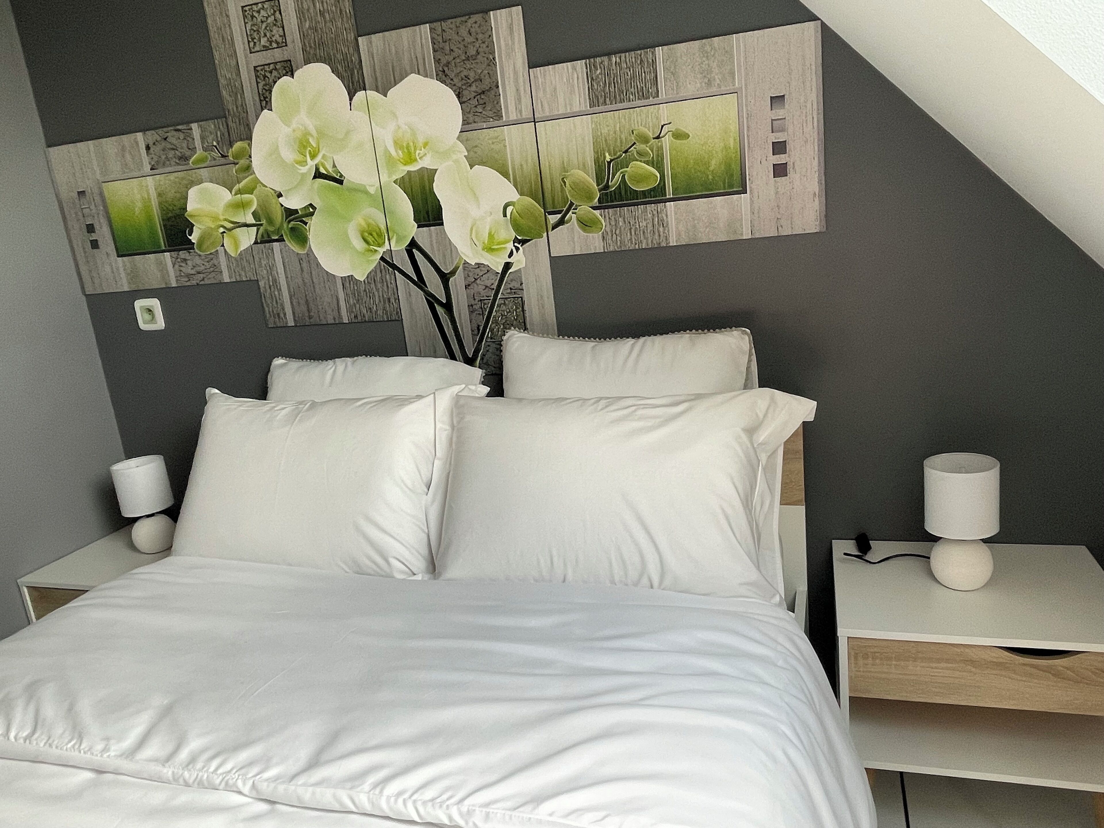 Un dormitorio moderno con cama, almohadas blancas, una mesita de noche, una lámpara y un florero con flores blancas.