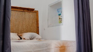 Un dormitorio con cama, cabecero tejido, pequeña ventana con un cuadro y cortina.