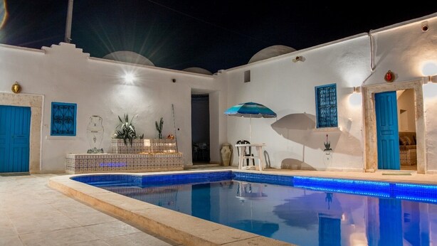 Un área junto a la piscina con una pileta con azulejos, una zona de estar y una mesa pequeña con un paraguas azul.