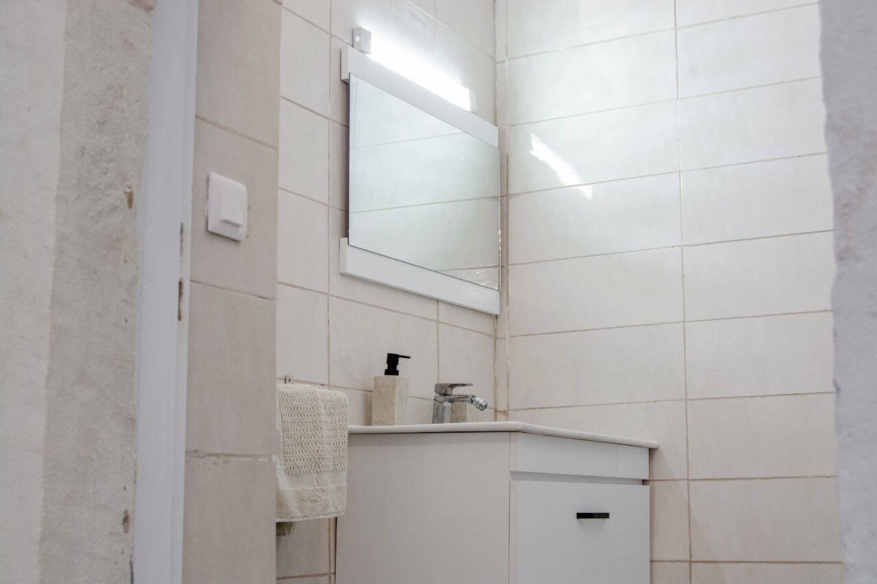Un bagno con piastrelle bianche, uno specchio, un lavandino con dispenser di sapone e un asciugamano appeso al muro.