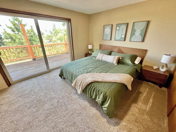 A bedroom with a large bed, a nightstand, a lamp, and a view of a wooden deck through a sliding glass door.