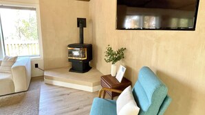Een gezellige woonkamer met een houten plafond, een inbouwvuurplaats, een comfortabele teal fauteuil en een groot raam met uitzicht op de omgeving.