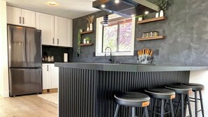 Een moderne keuken met een centraal eiland, barkrukken, een roestvrijstalen koelkast en een raam met uitzicht op bomen.