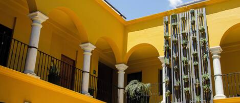 A yellow building with multiple arches and columns, a balcony with potted plants, and a staircase.
