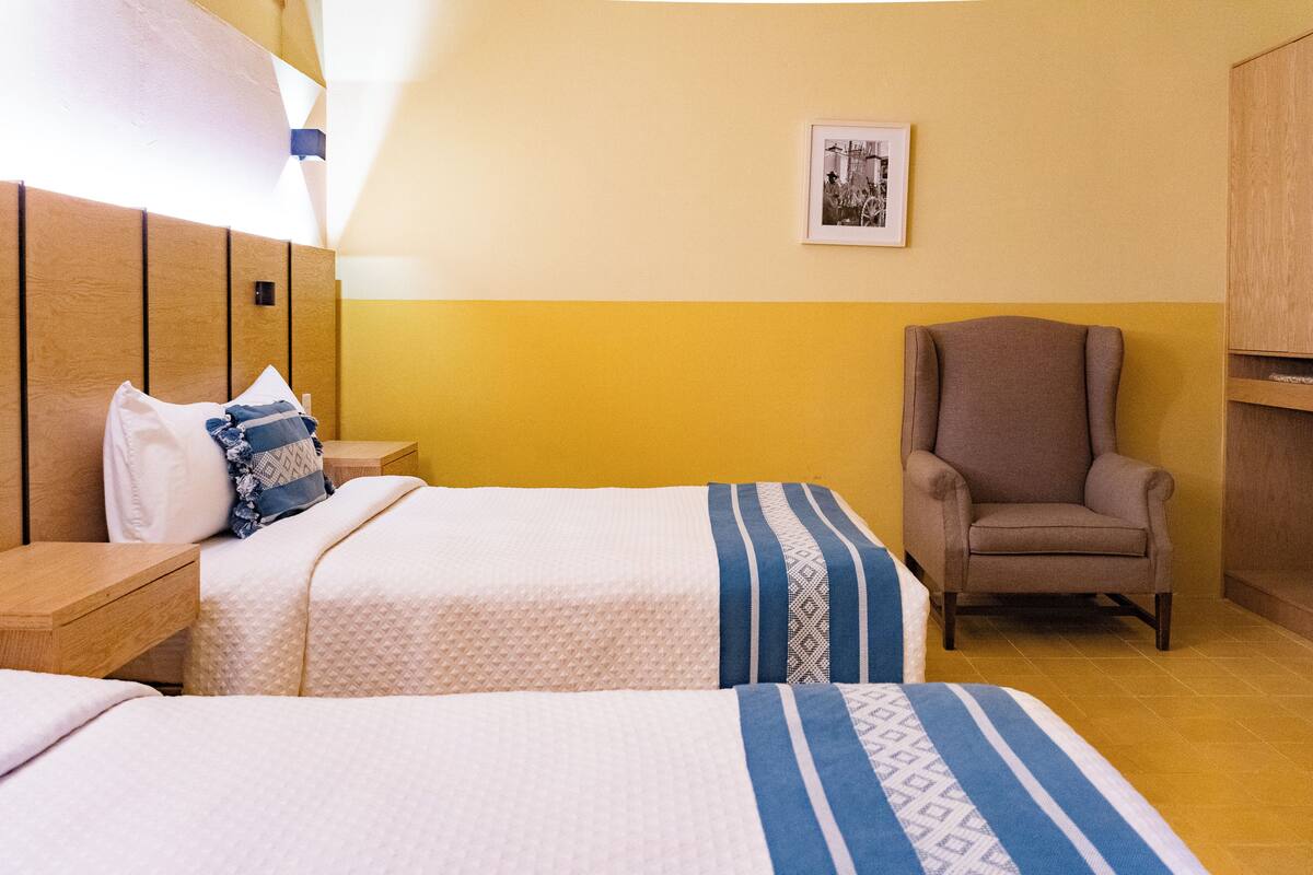 A hotel room with two beds, a chair, a wooden headboard, a framed picture on the wall, and a yellow and white color scheme.