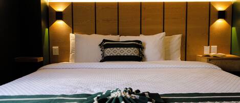 A neatly made bed with white bedding and a green and white striped blanket, wooden headboard, and two wall-mounted nightstands with lamps.