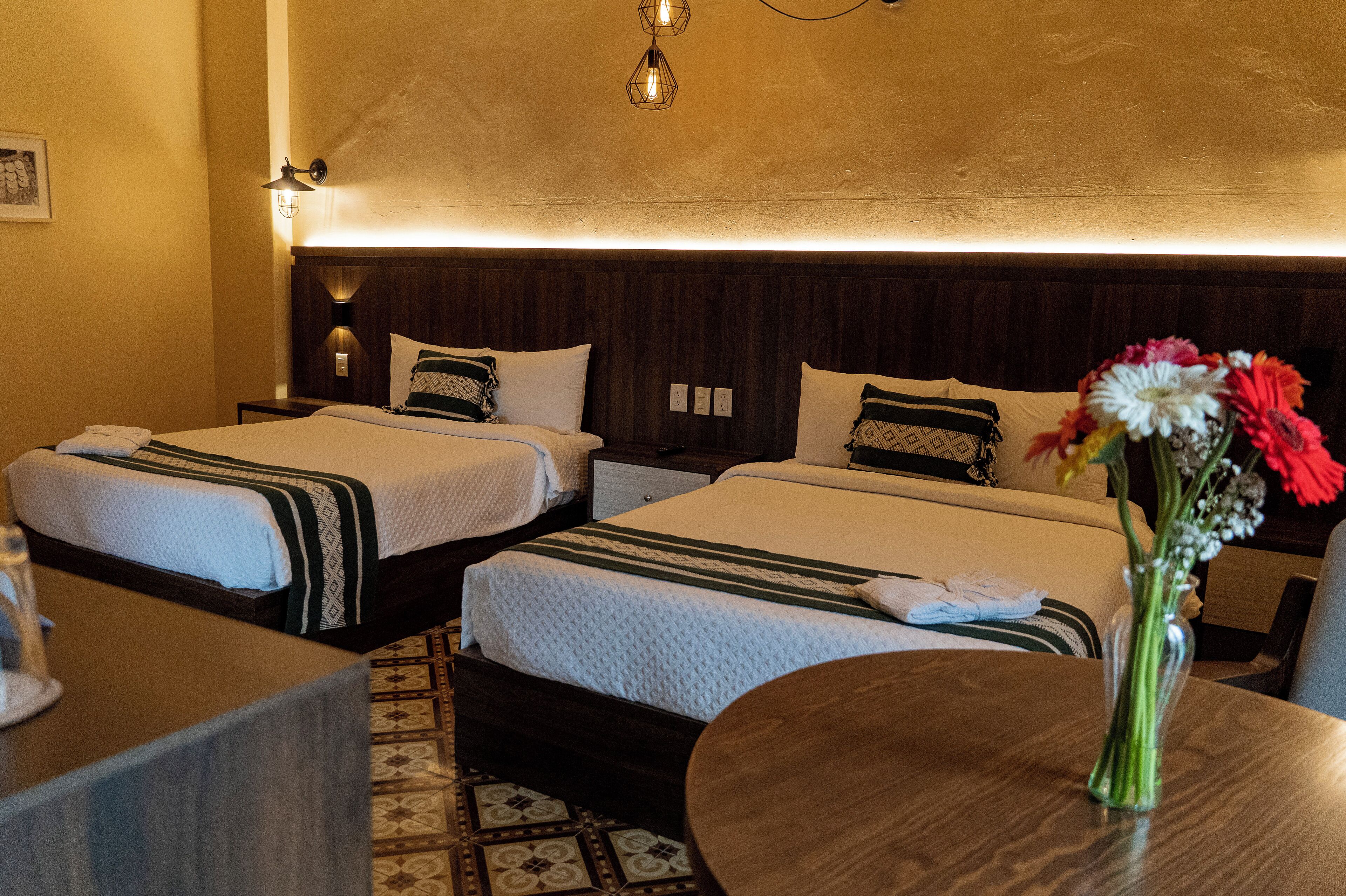 Habitación de hotel con dos camas, un cabecero de madera, una mesa redonda y un florero con flores.