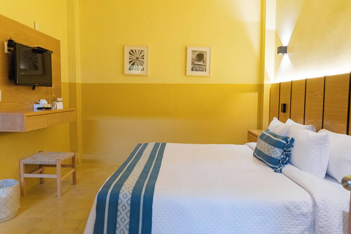 A hotel room with a bed, a small stool, a wooden nightstand, a wall-mounted TV, and two framed pictures on the wall.