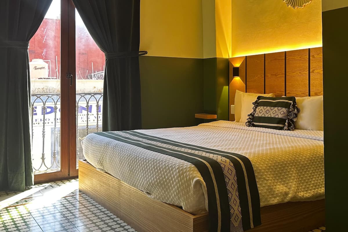 A hotel room with a bed, a nightstand, a lamp, and a view of the outside.