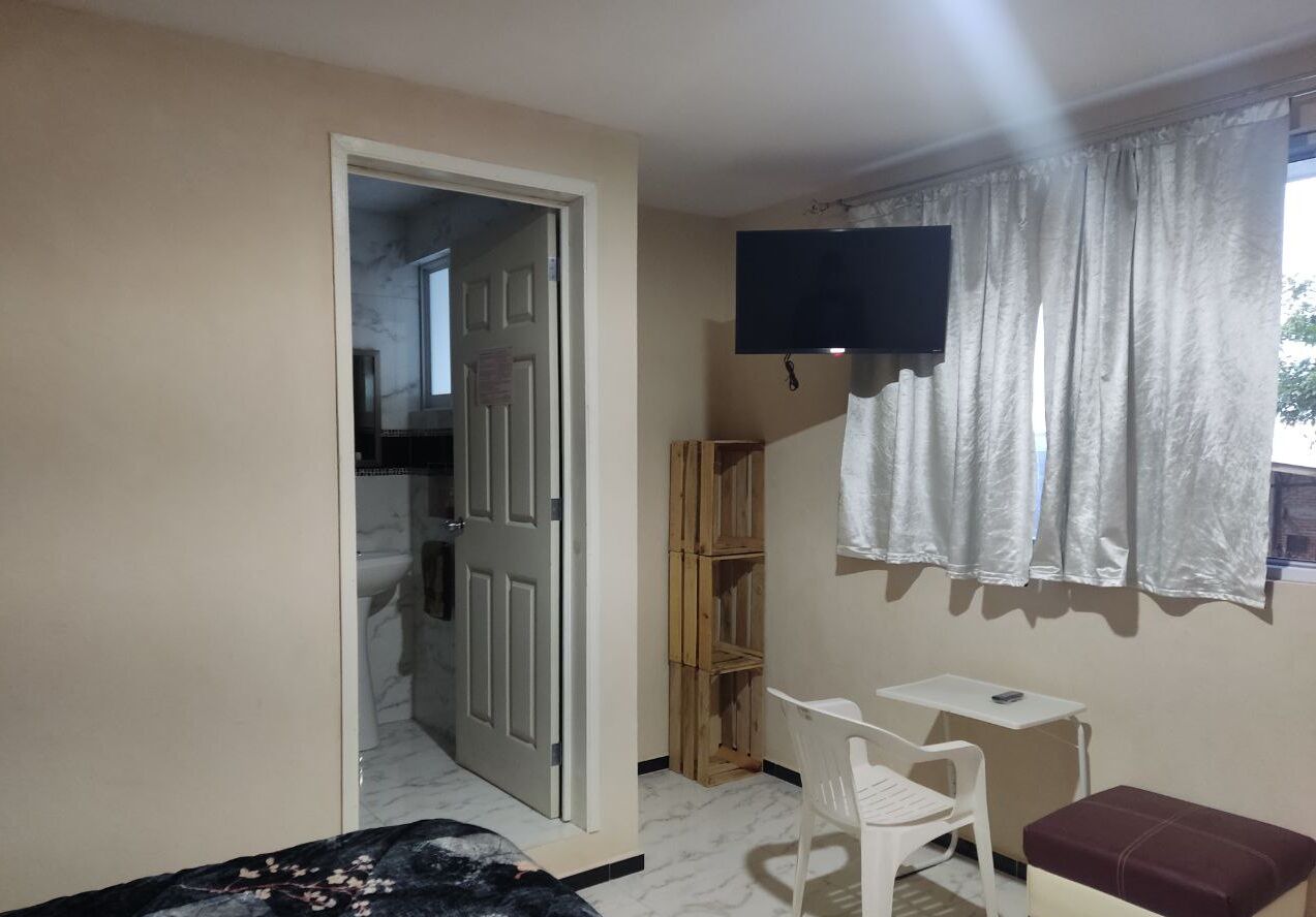 Una habitación pequeña con cama, escritorio, silla, televisor montado en la pared y ventana con cortinas.