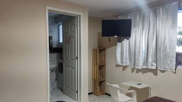Una habitación pequeña con cama, escritorio, silla, televisor montado en la pared y ventana con cortinas.