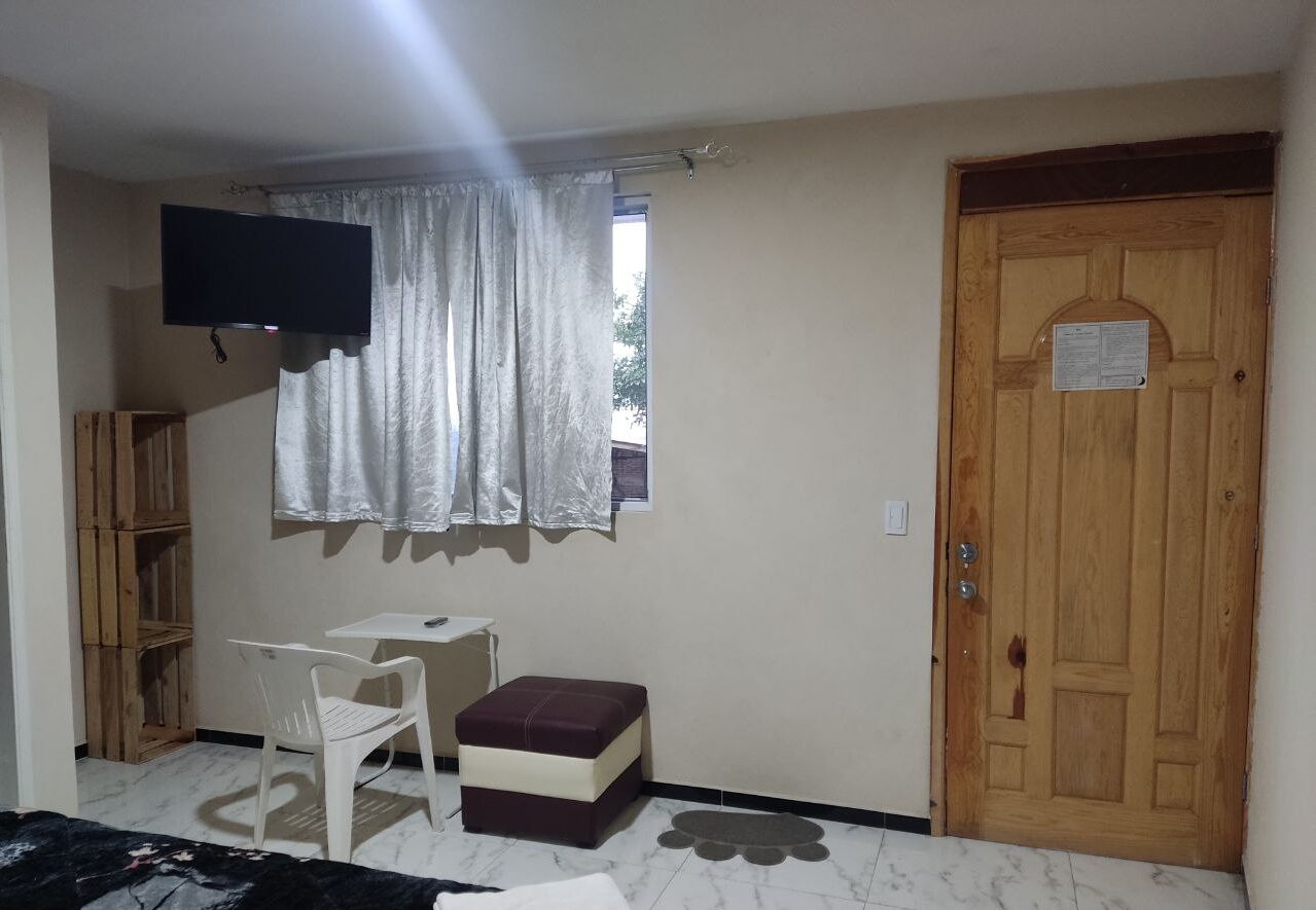 A small room with a television, a window with curtains, a wooden door, a small table, a chair, and a bench.