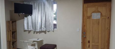 A small room with a television, a window with curtains, a wooden door, a small table, a chair, and a bench.