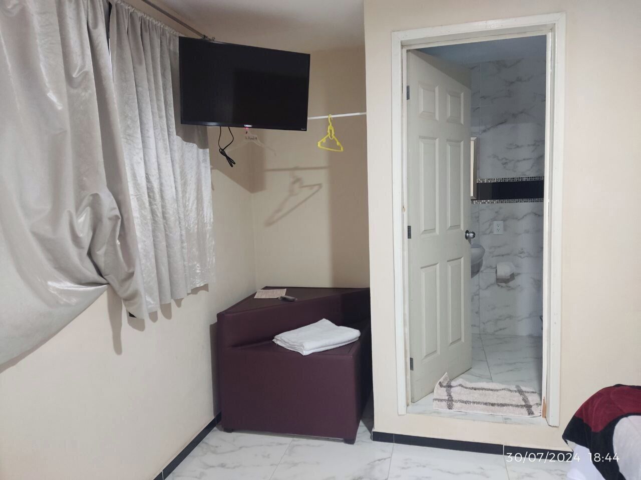 Una habitación pequeña con un televisor montado en la pared, un sofá de color oscuro, una puerta que da a un baño y una cortina entreabierta.