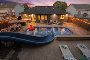 Un patio trasero con piscina, tobogán y mobiliario exterior.