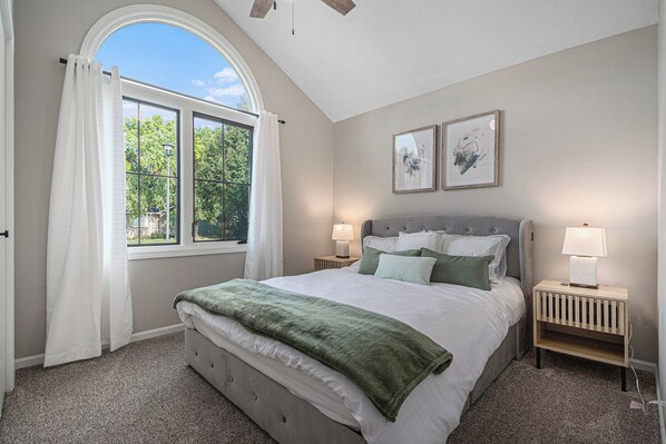 A bedroom with a large bed, two bedside tables with lamps, a window with curtains, and two framed pictures on the wall.
