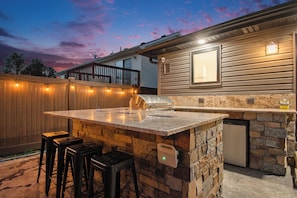 Una cocina al aire libre con una isla de piedra, taburetes de bar y un refrigerador empotrado.