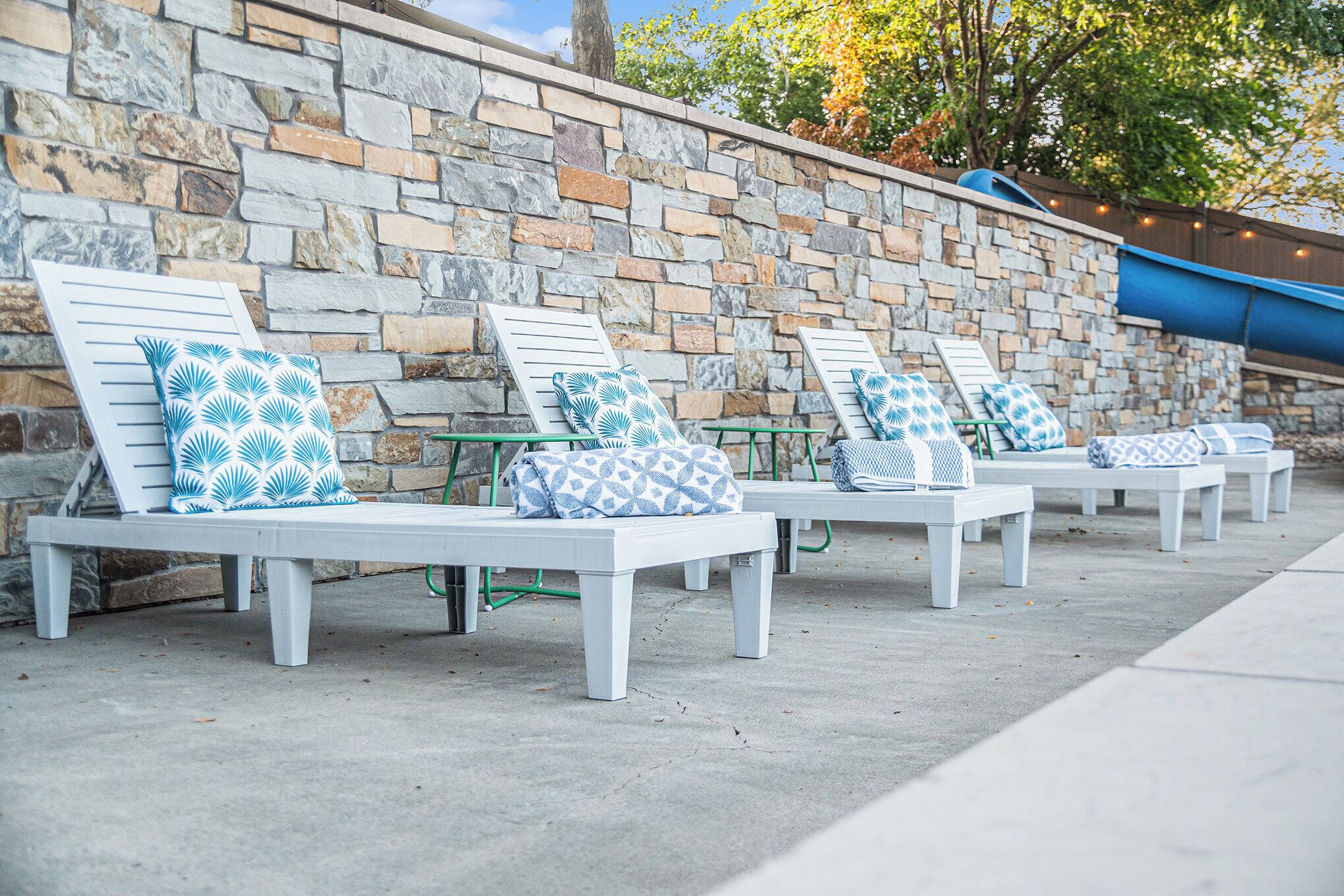 Outdoor lounge chairs with patterned cushions against a stone wall, with a water slide in the background.