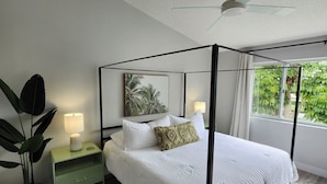 A modern bedroom with a four-poster bed, a ceiling fan, a nightstand with a lamp, and a large window with a view of greenery.