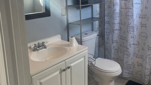 Ein Badezimmer mit einem weißen Waschbecken, Waschtisch und einer Toilette. Darüber befindet sich ein Ablagekorb aus Metall und eine Duschvorhang mit Aufschrift.